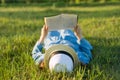 Girl in dress and hat lies on green grass reading book. View from above