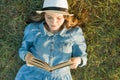 Girl in dress and hat lies on green grass reading book. View from above