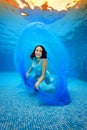 The girl in the dress posing underwater at the bottom of the pool, playing with a blue cloth, looking at the camera and smiling Royalty Free Stock Photo