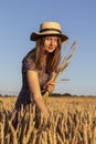 A girl in a dress and a straw hat collects spikelets in a wheat field
