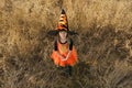A girl dressed as a witch in a huge black hat with an orange ribbon looks up standing in the tall dry grass. Royalty Free Stock Photo