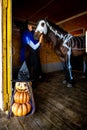 A girl dressed as a witch looks at a horse on which a skeleton is painted in white paint, in the foreground is an evil figurine of