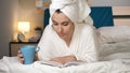 Girl drinking coffee or tea and reading book. Attractive woman in bedroom in white bathrobe with towel on her head lies on stomach Royalty Free Stock Photo