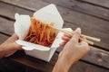 Girl eating wok noodles with vegetables and seafood in carton box to go, with bamboo chopsticks, closeup. Traditional Asian