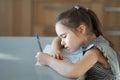 A girl of European appearance in a plaid dress is doing homework for the school at the table in the kitchen. The student