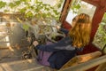 Girl with flowing blond hair in sunglasses sits in the cab at the driver`s seat in an abandoned ivy-covered bus