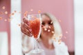 A girl in full focus holds a transparent glass of champagne, from which wax in the form of granules for depilation of pink color