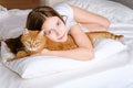 A girl with a ginger cat lies on a white bed. A young girl is stroking a ginger cat while lying on a white sheet