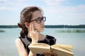 A girl with glasses reflects while reading a book sitting by the lake