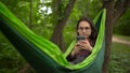 A girl in glasses swings in a hammock in nature and listens to music. A young woman puts wireless headphones into her Royalty Free Stock Photo