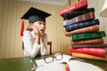 Girl in graduation cap looking at high heap of books on table at