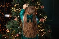 Girl in green dress and with a bowknot of red ribbon on her hair is standing in living room from behind near Christmas tree.