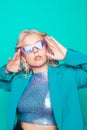 A girl in a green jacket and purple glasses poses on a green background. Fashion photo in 80s 90s style. Blonde girl in a shiny Royalty Free Stock Photo