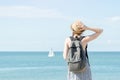 Girl in a hat with a backpack standing on the coastline. Sailboat in the distance. Back view