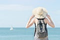Girl in a hat with a backpack standing on the coastline. Sailboat in the distance. Back view