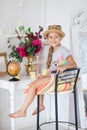 A girl in a hat sits on a high chair in a white room with a small globe, a little traveler