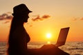 Girl in a hat sitting and working at his laptop against the sea at sunset. Silhouette Royalty Free Stock Photo