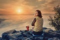 Girl is having breakfast and drinking coffee on a cliff top with a view of sunrise in mountains, and a thick fog is creeping Royalty Free Stock Photo