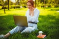 girl in headphones sits on the grass in the park with laptop talks through a video call with someone Royalty Free Stock Photo