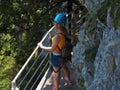 A girl in a helmet stands holding on to the railing on a narrow path of a rocky cliff in a green grove