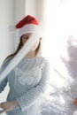 Girl hid behind a translucent curtain, in a New Year`s cap, next to a white artificial Christmas tree, close-up, copy space Royalty Free Stock Photo