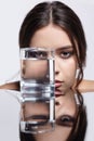 Girl hides her face behind a glass with water. Beauty portrait of young woman at the mirror table Royalty Free Stock Photo