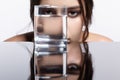Girl hides her face behind a glass with water. Beauty portrait of young woman at the mirror table Royalty Free Stock Photo