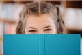 Girl hiding face behind a book with bookshelf in background Royalty Free Stock Photo