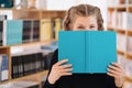Girl hiding face behind a book with bookshelf in background Royalty Free Stock Photo