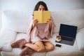 Girl hiding her face behind a book while sitting on the sofa with a laptop and yogurt or ice cream with fresh fruits in a jar Royalty Free Stock Photo