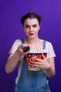 Girl holding large popcorn box, staring change channel while watching television with remote control isolated at purple Royalty Free Stock Photo