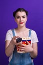 Girl holding large popcorn box, staring change channel while watching television with remote control isolated at purple Royalty Free Stock Photo