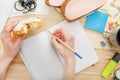 The girl is holding a pencil and a donut in her hands. On the wooden table there is a notebook, sandwiches...