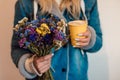 A girl holds flowers and a paper cup with coffee in her hands