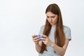 Girl holds smartphone in boths hands, looks confused at mobile screen, standing against white background