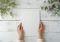 Girl holds a white poster mockup with no frame in her hands with green plants around on white wooden background Royalty Free Stock Photo