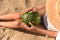 Girl holds a whole ripe green watermelon in her hands. picnic on the beach. young woman in straw hat with long legs sit on sand in Royalty Free Stock Photo