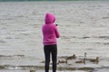 A girl in a hood stands with her back and takes pictures on her smartphone of ducklings and a duck floating by on the river