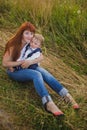 The girl hugs baby tightly, they are sitting on the grass, mom and son are smiling and happy, mom has red hair