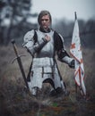 Girl in image of Jeanne d`Arc in armor kneels with flag in her hands and sword on meadow.