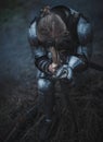 Girl in image of Jeanne d`Arc in armor and with sword in her hands kneels against background of dry grass.