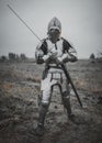 Girl in image of Jeanne d`Arc goes on meadow in armor and helmet with closed visor and sword in her hands.