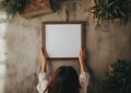 Girl inserts a white poster mockup into wooden frame with her hands, green plants around on gray background Royalty Free Stock Photo