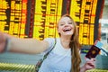 Girl in international airport, taking funny selfie with passport and boarding pass near flight information board Royalty Free Stock Photo