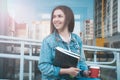 The girl in jeans clothes sits on a bench, holds notebooks and coffee. The girl smiles, studies in the open air and drinks coffee