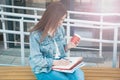 A girl in jeans clothes sits on a bench, holds notebooks, coffee and writes. The girl smiles, studies in the open air and drinks