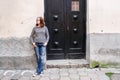 A girl in jeans, a striped sweater and sneakers is standing near the black wooden door of an old house and looks to the side Royalty Free Stock Photo