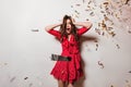Girl in joyful shock poses against background of confetti. Portrait of astonished dark-haired model in red dress.