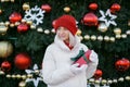girl in a knitted red hat and mittens holds a gift box in her hands while standing on the street by a decorated Christmas tree. Royalty Free Stock Photo
