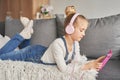 Girl laying on couch in headphones, listening to music with her smarthphone. Little girl watching cartoons on her phone. Listening